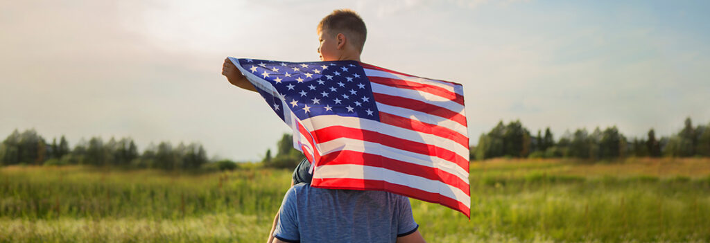 Photo of a boy holding an American flag while sitting on his father's shoulders in a grassy field.